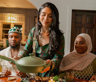 A woman in a green dress serves food from a green pot at a dining table, with two people sitting beside her, one wearing a kufi and the other wearing a hijab.