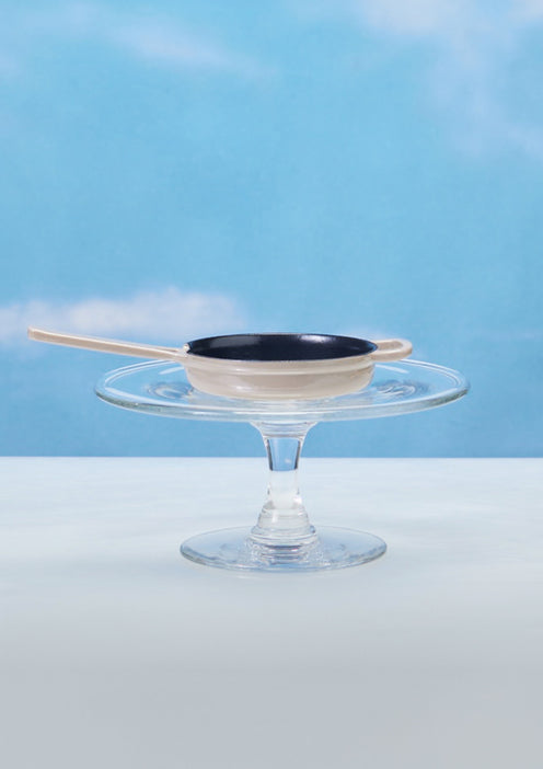 A small black pan with a wooden handle rests on a glass cake stand against a blue sky background.
