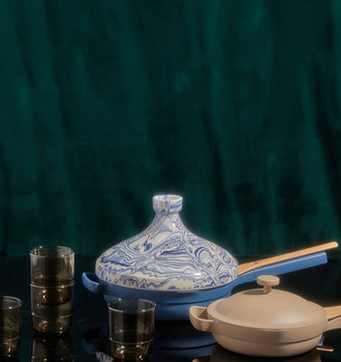 Assorted tableware including stacked plates, bowls, glasses, a green pot, a beige pot, and a blue-patterned tagine dish displayed on a reflective surface with a dark green backdrop.