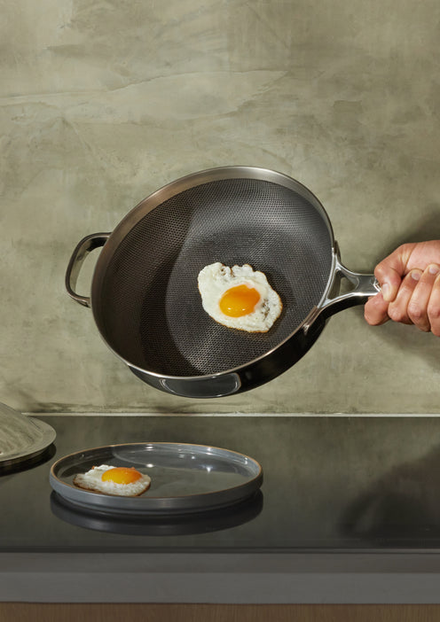 A hand holds a frying pan with a cooked sunny-side-up egg as it is being placed onto a metal plate on a kitchen countertop.