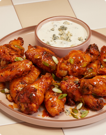 A plate of buffalo chicken wings garnished with sliced green onions, served with a bowl of blue cheese dip on a tiled surface.