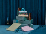 A stack of three teal gift boxes with blue bows, an envelope, a pen, two amber glasses, a pink oven mitt, and a card reading Lets dig in on a blue velvet surface and background.