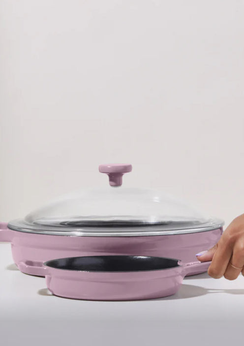 A hand holds a small pink frying pan in front of a larger pink pan with a glass lid on a white surface.