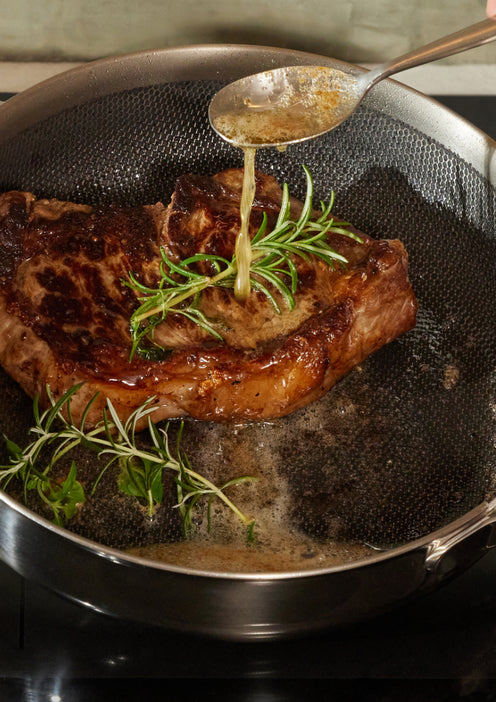 A cooked steak in a frying pan is being basted with melted butter using a spoon, with fresh rosemary and thyme beside it.