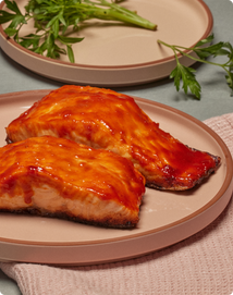 Two pieces of glazed salmon are served on a beige plate, with a pink cloth and a plate with parsley in the background.