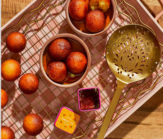 A tray with bowls of gulab jamun, a golden slotted spoon, scattered gulab jamun balls, and a small tin of saffron threads.
