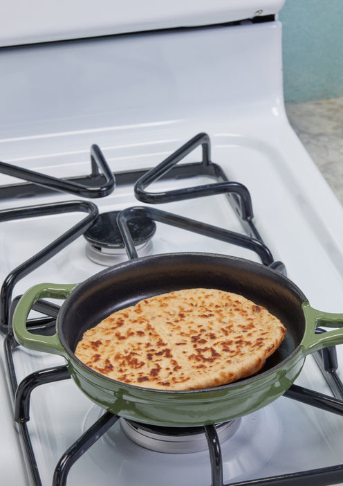 A round, golden-brown flatbread is cooking in a green-handled skillet on a white gas stove.