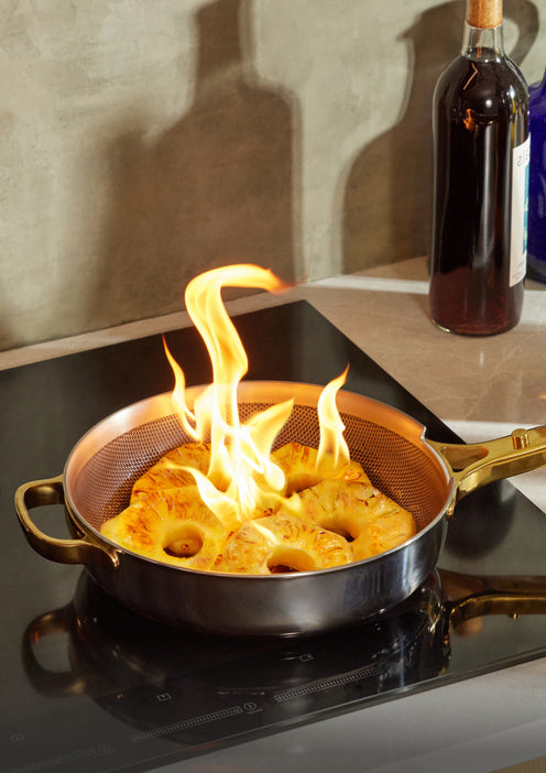 Four pineapple slices in a pan are being flambéed on a stovetop, with visible flames rising from the fruit. A bottle stands in the background on the counter.