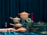 Assorted cookware in earthy tones is arranged on teal gift boxes with ribbons, against a dark blue curtain backdrop. A single pink candle is lit among the items.