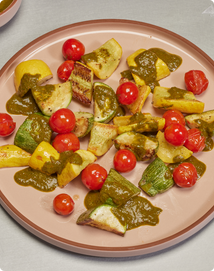 A round plate with roasted yellow squash, zucchini, and cherry tomatoes, topped with a green sauce, arranged on a light surface.