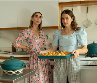 Two women stand in a kitchen; one holds a tray of baked rolls. Both wear patterned dresses and look toward the camera with neutral expressions.