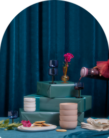 A table with stacked teal gift boxes, cookies on a plate, pink bowls, wine glasses, flowers in a vase, and a hand pouring wine, set against a blue velvet curtain backdrop.