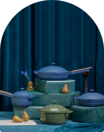 A set of blue and green cookware with wooden handles displayed on turquoise boxes, surrounded by pears and small vases, against a blue velvet curtain backdrop.