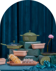 Assorted cookware, including green pots and pink baking dishes, arranged on teal gift boxes with a ribbon, glass bundt mold, and a single pink flower against a dark teal curtain.