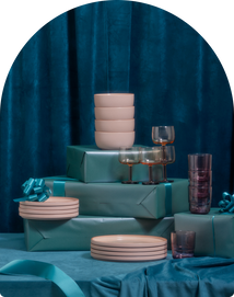 Stacks of pink dinnerware and various glassware are arranged on teal gift-wrapped boxes with blue bows, set against a blue velvet curtain background.