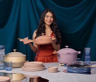 A woman in an orange dress holds a pink pan, standing behind a table arranged with various cookware, plates, and glasses against a blue draped background.