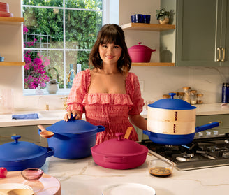 A woman in a pink dress stands in a kitchen with pink and blue pots and pans on a countertop and stove, with a window and shelves in the background.