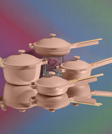 A set of four pink pots and pans with wooden handles and lids is arranged on a reflective surface, with a blurred multicolored background.