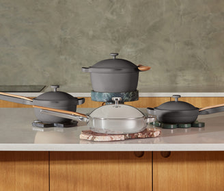 Four pieces of cookware, including saucepans and a pot with lids, are arranged on a kitchen counter with stone trivets.