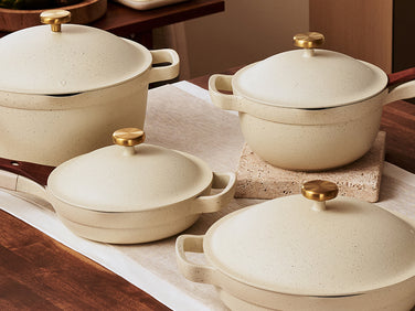 Four cream-colored ceramic pots and pans with wooden handles and gold knobs are arranged on a kitchen countertop.