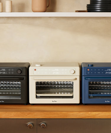 Three Our Place countertop ovens in black, cream, and blue lined up on a wooden counter.