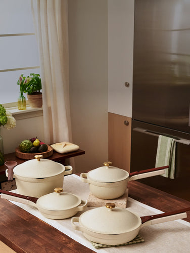 Four cream-colored ceramic pots and pans with wooden handles and gold knobs are arranged on a kitchen countertop.
