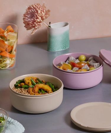 A table with two round containers of salad and fruit, a glass with layered vegetables, lids, and a ceramic cup with a pink flower against a light pink wall.