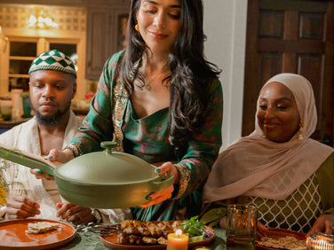 A woman in festive attire serves food from a green pan at a dining table with two other people, surrounded by plates of food, candles, and drinks.