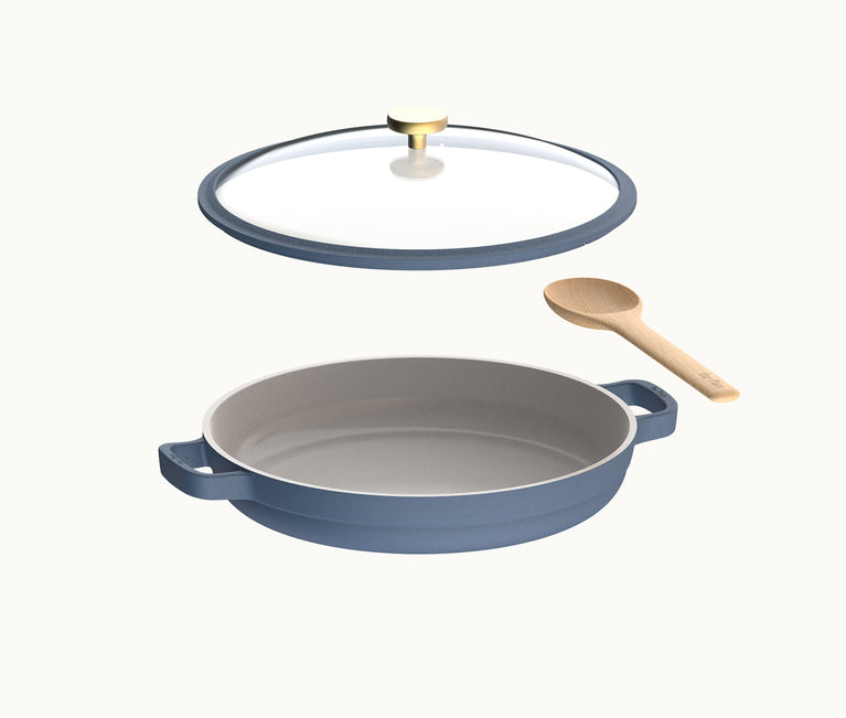 Blue ceramic pan with two handles, a glass lid with a brass knob, and a wooden spoon, all shown against a plain white background.