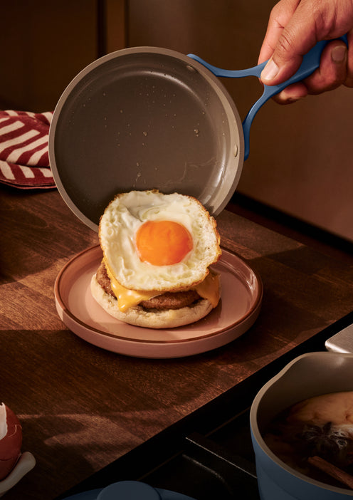 A hand places a fried egg from a pan onto a breakfast sandwich with cheese and sausage on an English muffin, set on a plate on a wooden countertop.