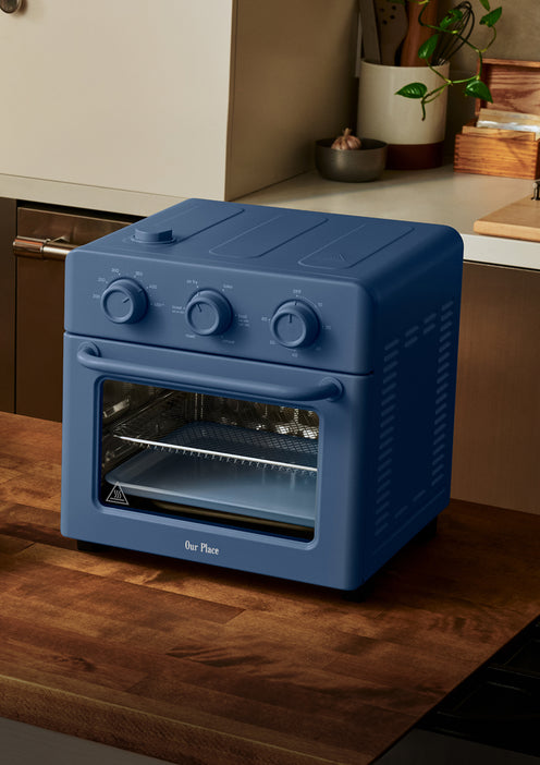 A blue countertop toaster oven with three control knobs sits on a wooden kitchen counter near a plant and utensils.