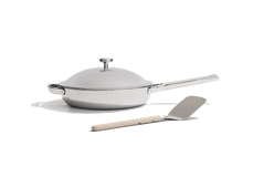 A stainless steel frying pan with a matching lid and a metal spatula with a light-colored handle on a white background.