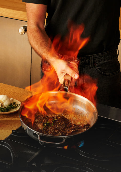 A person flambéing a steak in a stainless steel skillet on a stovetop.
