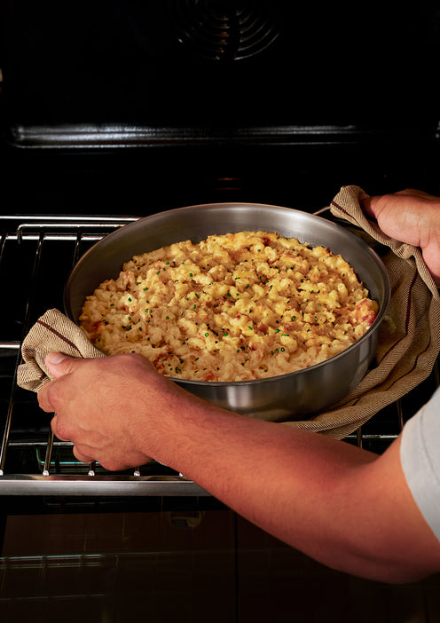 A person uses a towel to remove a round pan of baked macaroni and cheese topped with herbs from the oven.