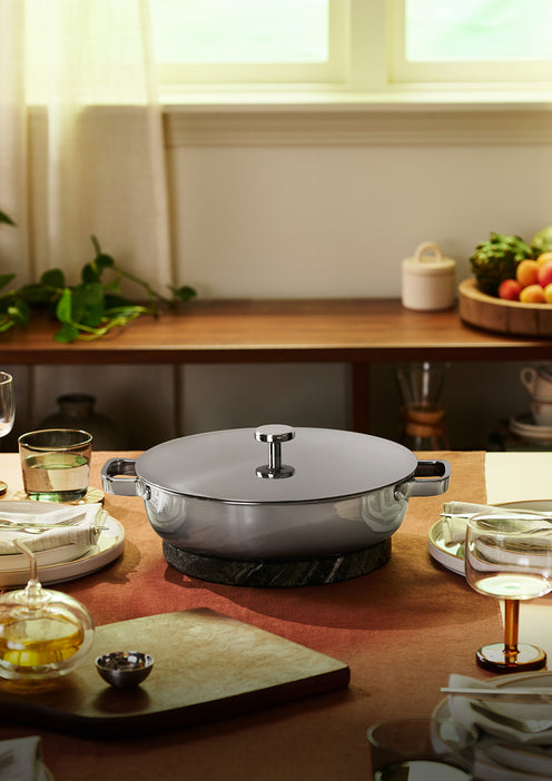 A lidded stainless steel pot sits on a trivet at the center of a set dining table with glassware, dishes, and food, in a sunlit room.