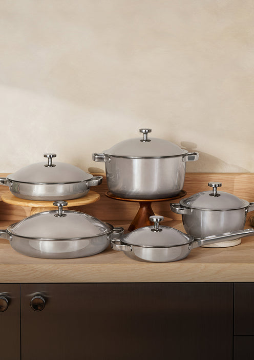 Five stainless steel pots and pans with lids are arranged neatly on a wooden countertop against a light beige wall.