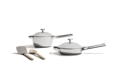 Two stainless steel pans with lids are placed next to a metal spatula and a wooden spoon on a white background.