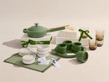 A green cookware set, cups, glasses, bowls, and serving items are arranged on and around white gift boxes with green and white ribbons on a beige background.