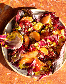 A plate of radicchio salad topped with orange and grapefruit segments, pomegranate seeds, chopped pistachios, and a yellow vinaigrette on an orange stone surface.