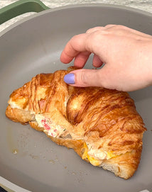 A hand with lavender nail polish holds a croissant filled with egg and other ingredients inside a gray frying pan.