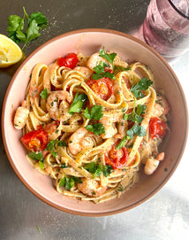 A bowl of fettuccine pasta with shrimp, cherry tomatoes, fresh parsley, and a creamy sauce, next to a lemon wedge and a glass of water.