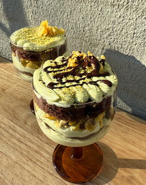 Two glass dessert cups filled with layered green cream, chocolate cake, and crunchy toppings, with chocolate drizzle on top, placed on a wooden surface in sunlight.