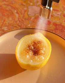 A lemon half is being caramelized with a culinary torch on a beige plate, with a mottled orange background.