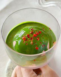 A glass dessert featuring a scoop of bright green matcha ice cream, topped with crushed pink peppercorns.