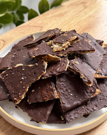 A plate filled with pieces of chocolate-covered toffee bark sprinkled with sea salt, set on a wooden surface with green leaves in the background.