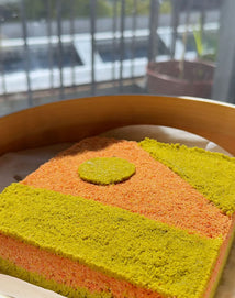 A colorful, square-shaped layered cake with orange and green sections and a green circle on top, placed in a round wooden container near a sunlit window.