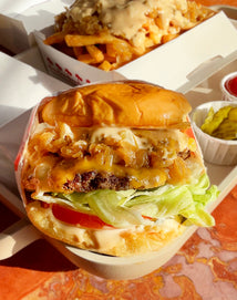 A close-up of a cheeseburger with lettuce, tomato, grilled onions, and sauce, with a side of pickles and fries topped with sauce in the background.