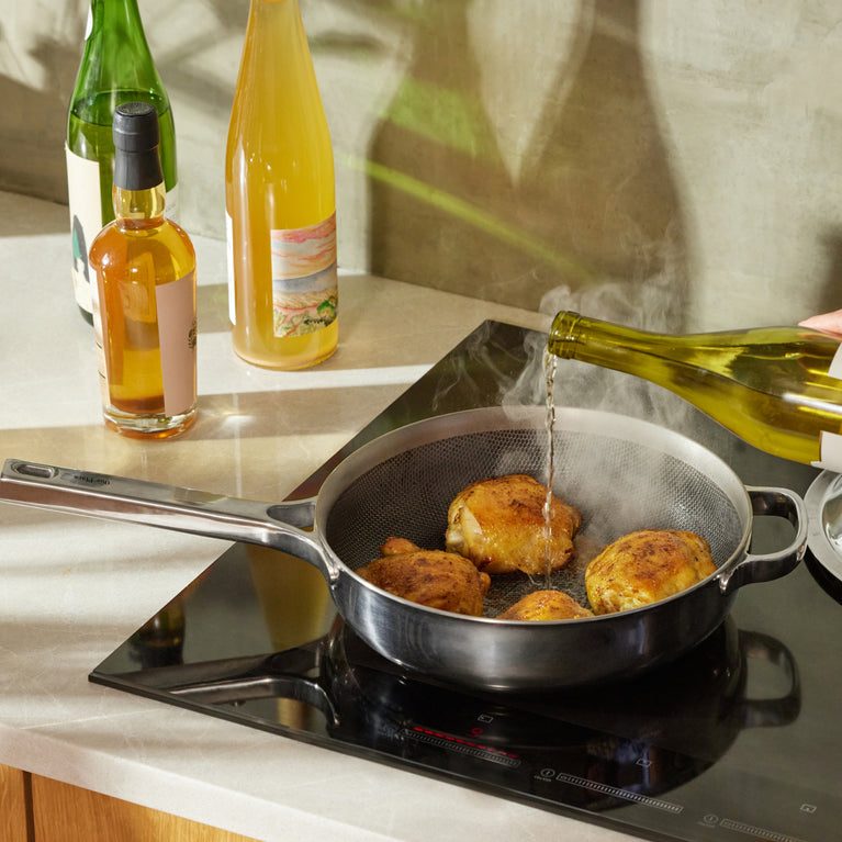 Four seasoned chicken thighs are cooking in a frying pan on an induction stove while wine is being poured into the pan; bottles are visible on the counter nearby.