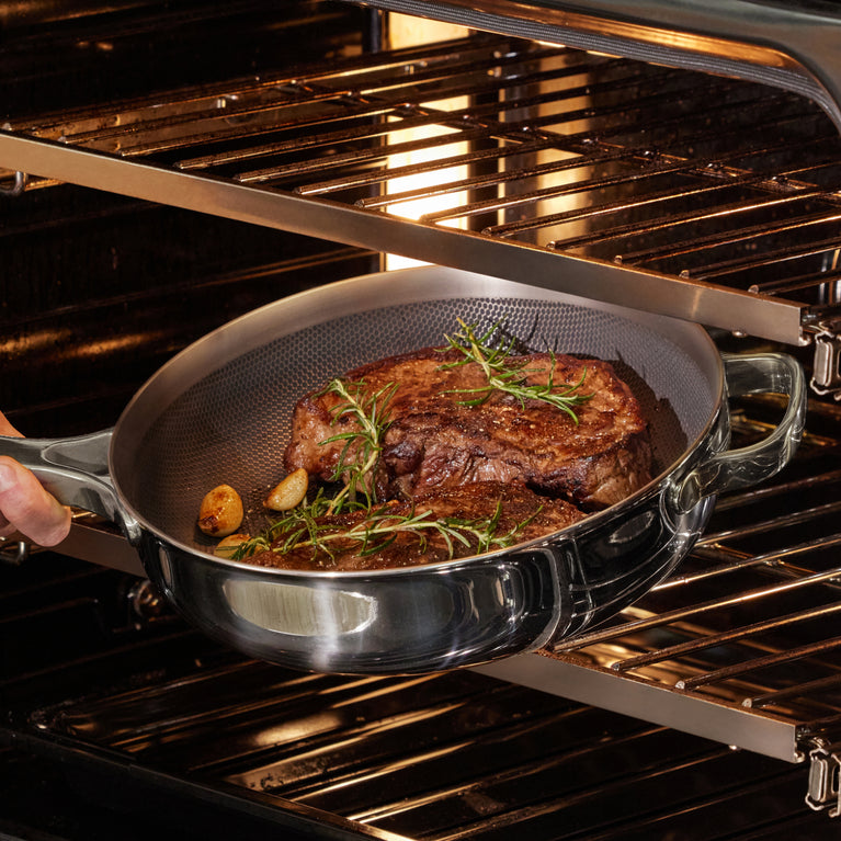 A person places a stainless steel pan with cooked steaks, garlic, and herbs into an oven with metal racks.