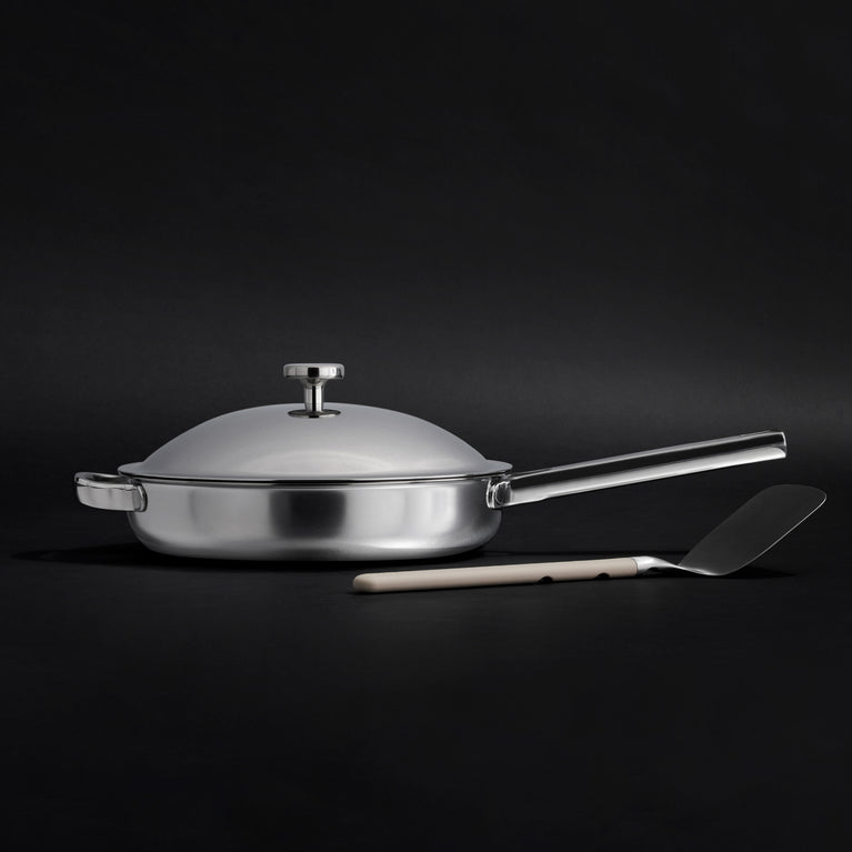 The Titanium Always Pan Pro, a white frying pan with a metal handle and advanced nonstick technology, sits beside a beige-handled spatula on a light background.